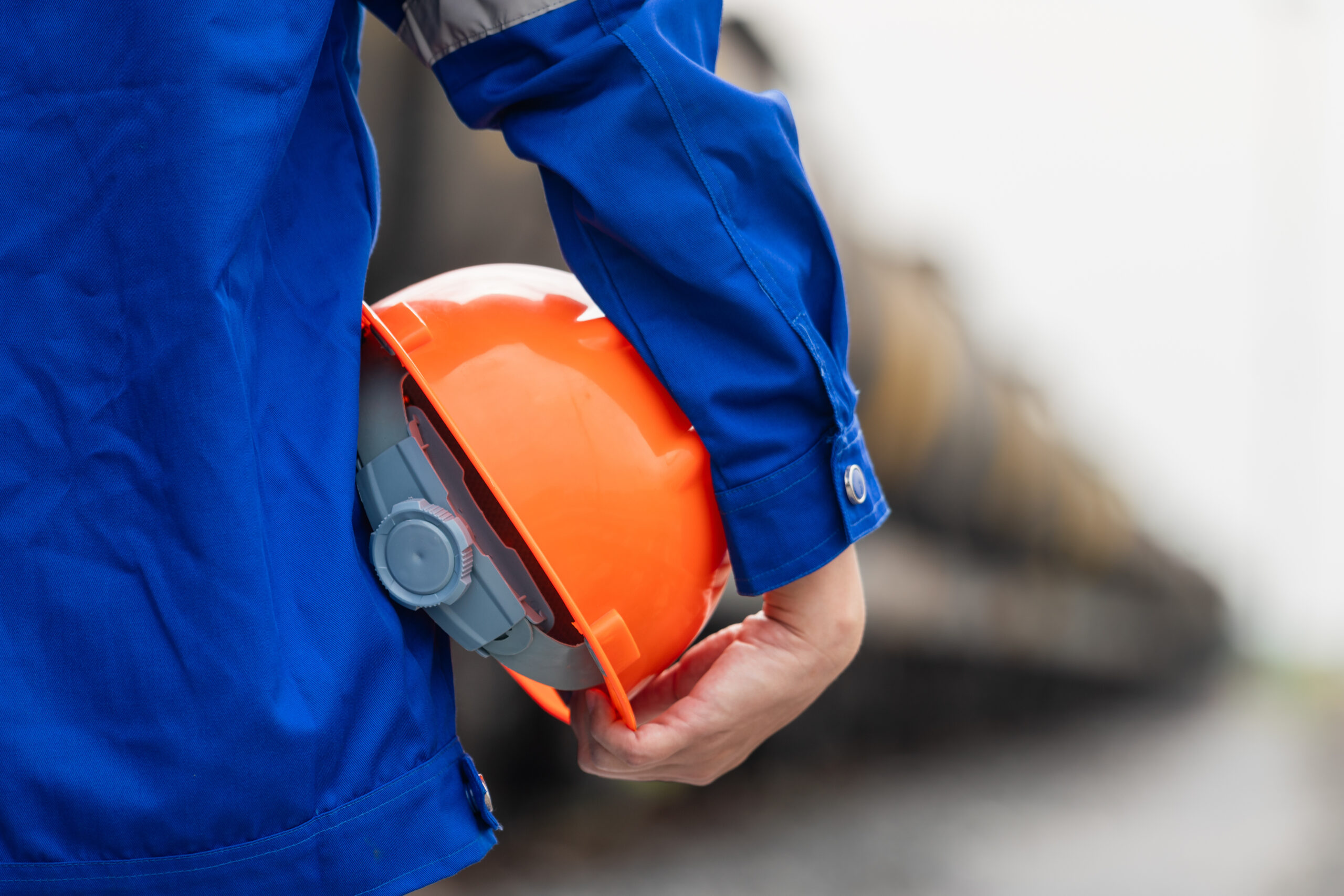 A worker in blue coverall uniform is holding an orange safety hardhat or protective helmet, standing in front of crude oil train tanker. Ready to work in challenge industrial scene, close-up.